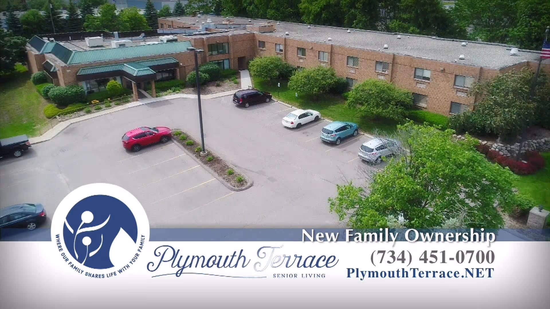 Aerial view of Plymouth Terrace senior living facility showing a single-story brick building with a green roof, surrounded by trees and shrubs. Several cars are parked in the parking lot in front of the building.