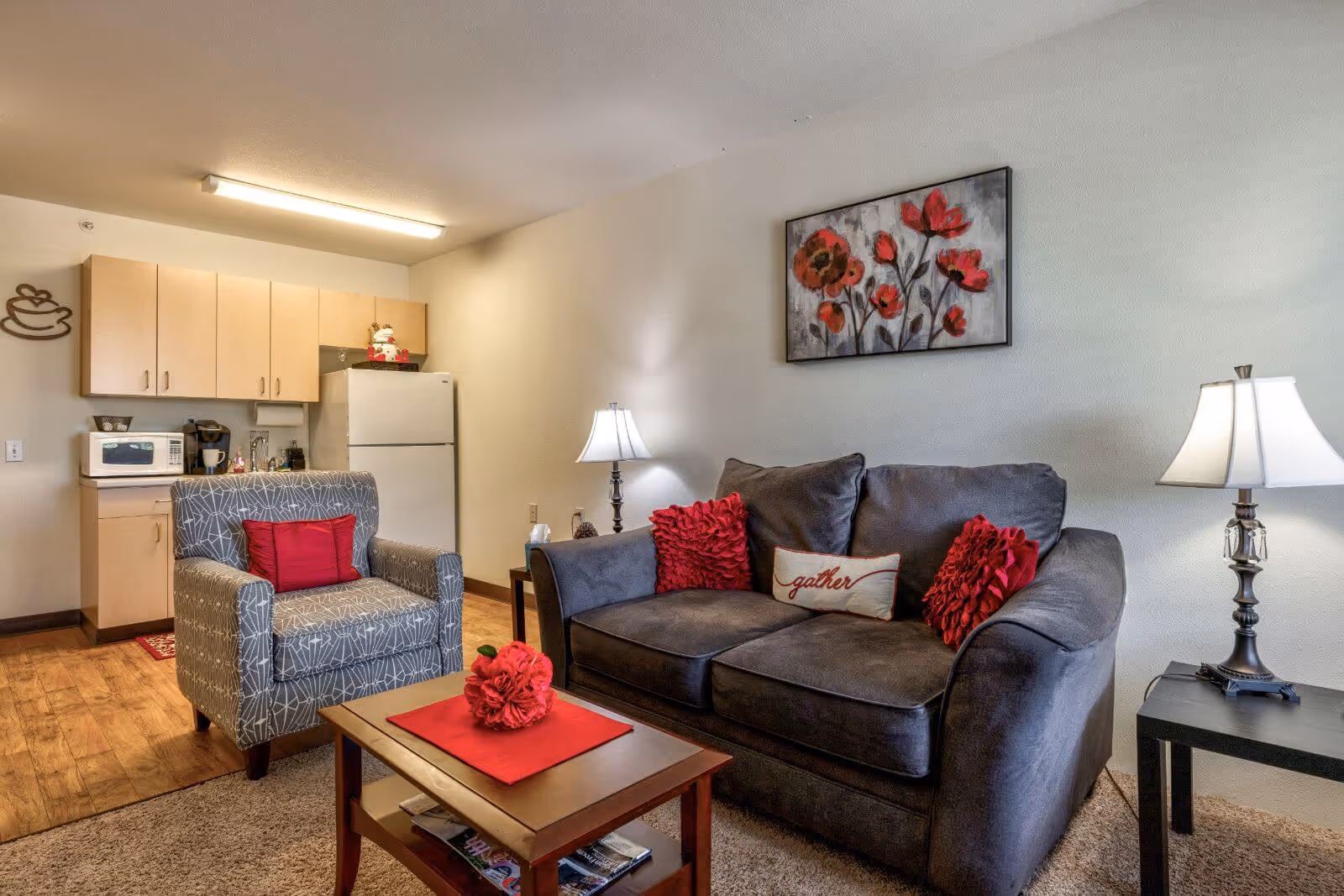 A cozy living room area with a dark gray couch adorned with red and white decorative pillows, a gray patterned armchair with a red pillow, a wooden coffee table with a red centerpiece, and two side tables with lamps. In the background, there is a small kitchen area with light wood cabinets, a white refrigerator, a microwave, and a coffee maker. A painting of red flowers hangs on the wall above the couch.