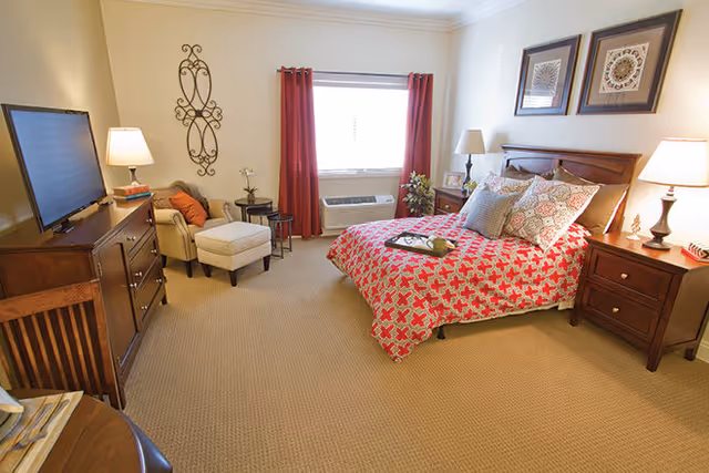 A cozy bedroom with a bed covered in a red and white patterned quilt, multiple pillows, two framed artworks above the headboard, a wooden nightstand with a lamp, a cushioned armchair with an ottoman, a wooden dresser with a flat-screen TV, and a window with red curtains letting in natural light.