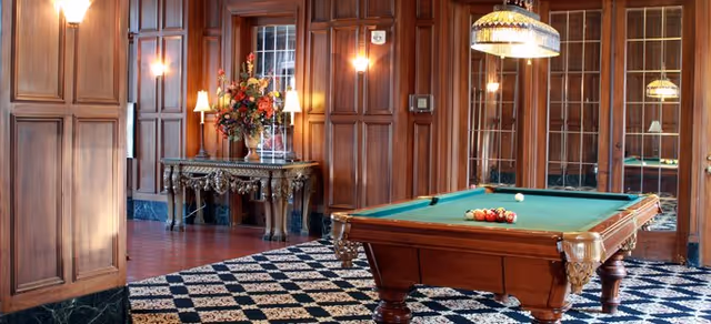 An elegant interior room featuring a wooden pool table with billiard balls arranged on the green felt surface. The room has rich wood-paneled walls, a patterned carpet, a decorative table with a floral arrangement, and warm lighting from wall sconces and a hanging lamp above the pool table. Large windows with wooden frames are visible in the background.