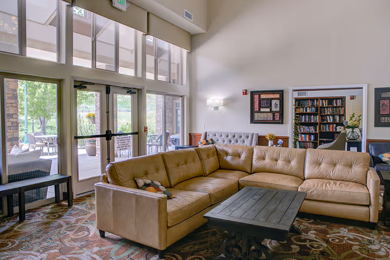 A spacious senior living facility common area with a large beige sectional sofa and a dark wooden coffee table on a patterned carpet. The room has high ceilings with large windows and glass doors leading to an outdoor patio with seating. There are bookshelves filled with books and framed artwork on the walls.