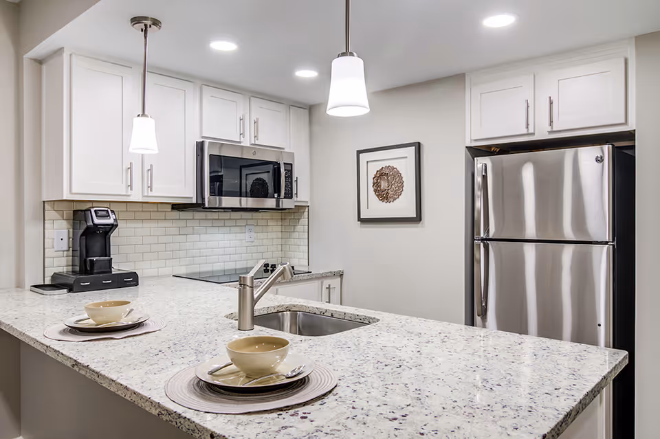 Modern kitchen with white cabinets, a stainless steel refrigerator, microwave, and a coffee maker. The kitchen features a granite countertop with an inset sink and two place settings with bowls, plates, and spoons. Two pendant lights hang from the ceiling, and a framed decorative artwork is on the wall.