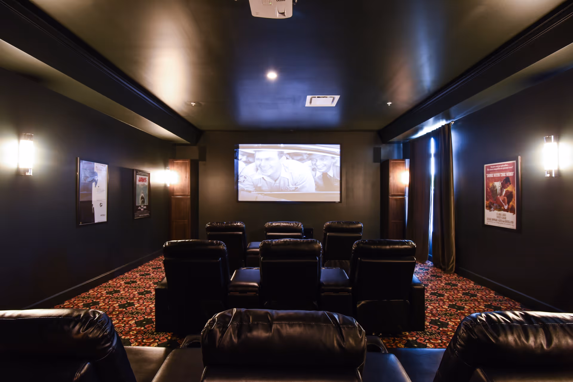 A cozy home theater room with black leather recliner chairs arranged in rows facing a large screen showing a black and white movie. The room has dark walls, patterned carpet, wall-mounted lights, and movie posters on the walls.