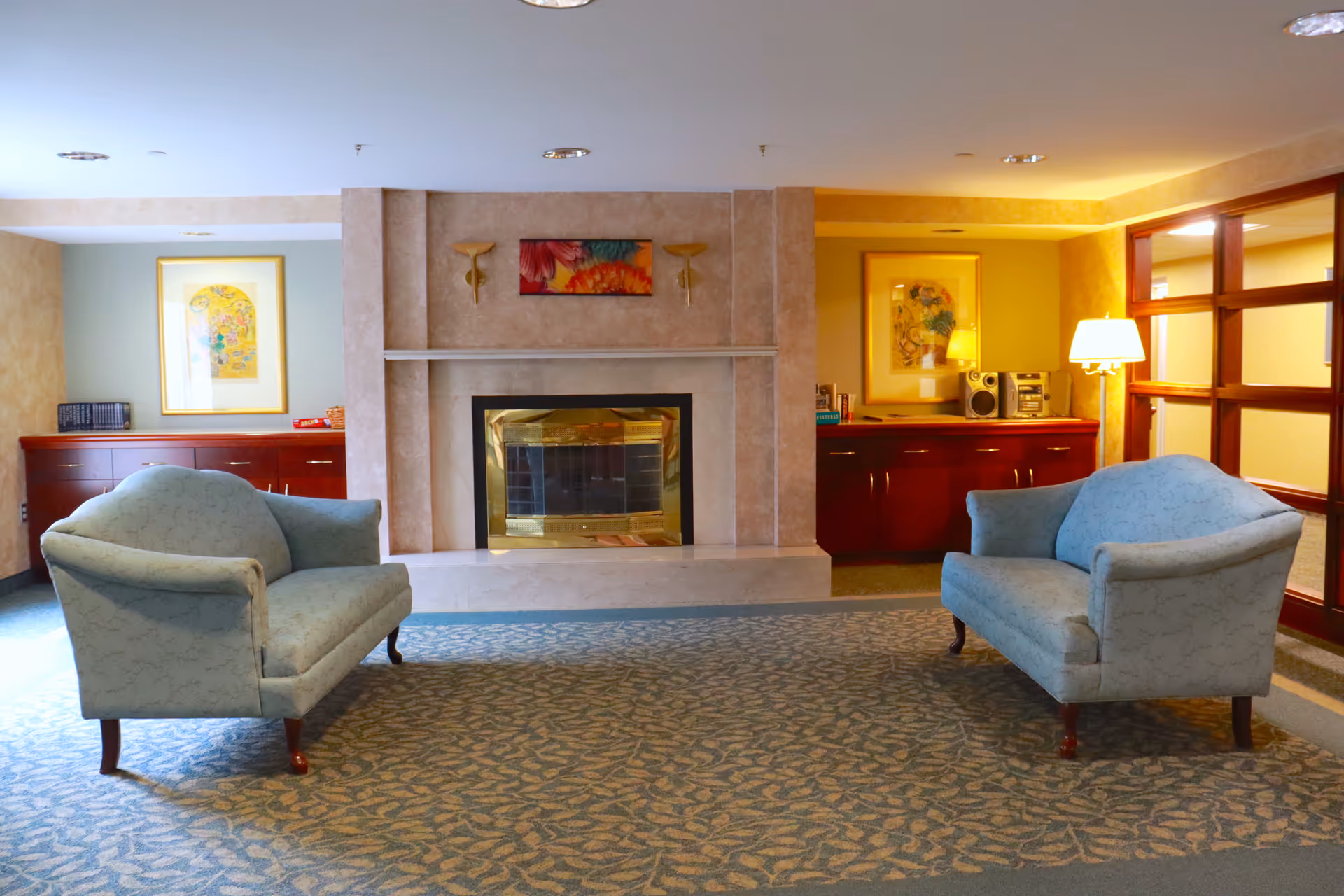 A cozy living room area in an assisted living facility featuring two light blue upholstered loveseats facing each other on a patterned carpet. In the center background is a fireplace with a beige stone surround and a colorful floral painting above it. On either side of the fireplace are wooden cabinets with framed artwork and a stereo system on the right. A floor lamp is also visible near the right loveseat.