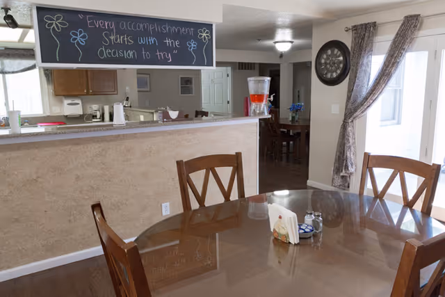Dining area with a glass-top table and wooden chairs. In the background, there is a kitchen counter with a chalkboard sign above it that reads 'Every accomplishment starts with the decision to try'. The room has light-colored walls, a clock on the wall, and a window with curtains letting in natural light.
