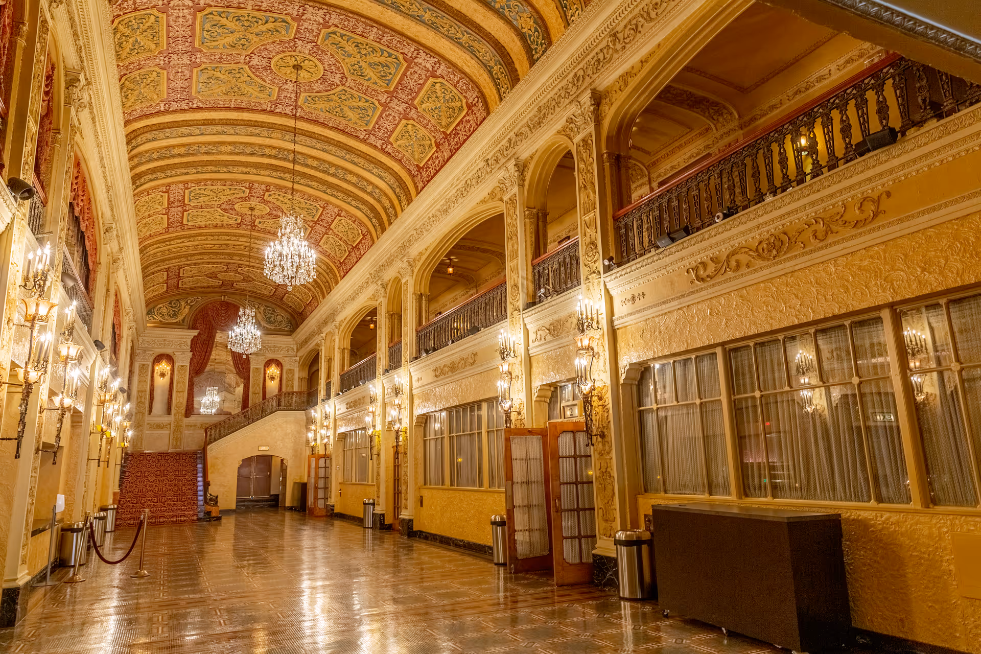 An ornate, grand interior hallway with a high, elaborately decorated ceiling featuring intricate patterns and chandeliers. The walls are adorned with detailed moldings and sconces with lights. There is a staircase at the far end, and large windows with curtains line one side of the hallway. The floor is polished and reflective.