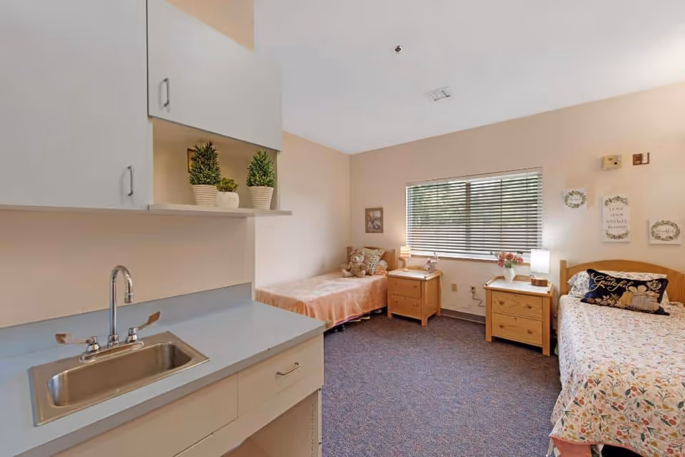 Shared resident bedroom with two single beds, nightstands, a window with blinds, and a small sink/cabinet area.