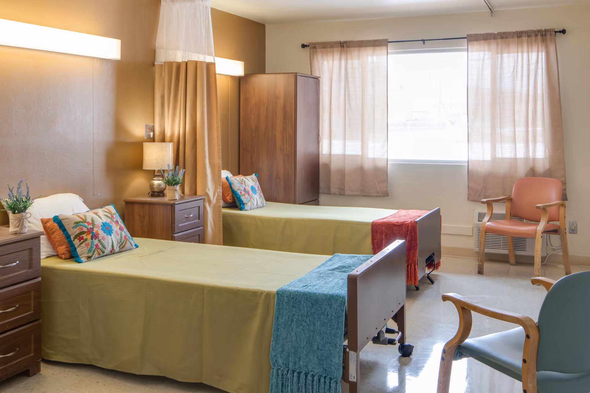 A bright and clean bedroom in a care facility with two single beds covered in light green bedspreads, each adorned with colorful pillows and a throw blanket at the foot. Between the beds is a wooden nightstand with a lamp and a small plant. A privacy curtain hangs between the beds. The room has a large window with beige curtains, a wooden wardrobe, and two chairs with wooden arms placed near the window.