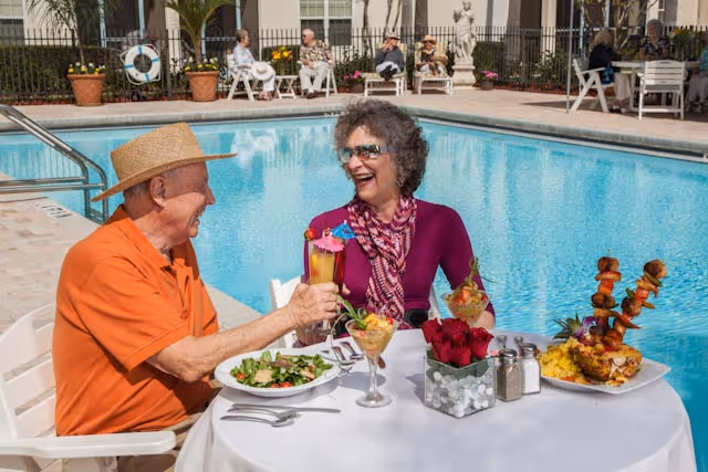 An elderly man and woman sitting at a table by a swimming pool, enjoying a meal and drinks. The man is wearing an orange shirt and straw hat, and the woman is wearing a purple top and scarf with sunglasses. The table is set with plates of food, drinks, and a centerpiece of red roses. In the background, other elderly people are seated near the pool.