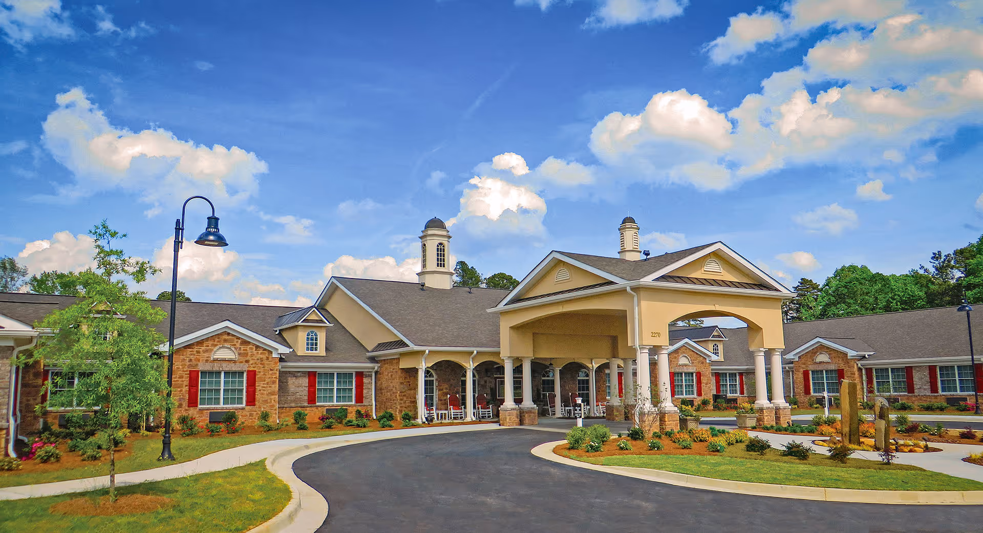 Front exterior view of Benton House of Grayson, a single-story building with a covered entrance supported by columns, red window shutters, and a landscaped circular driveway under a partly cloudy blue sky.