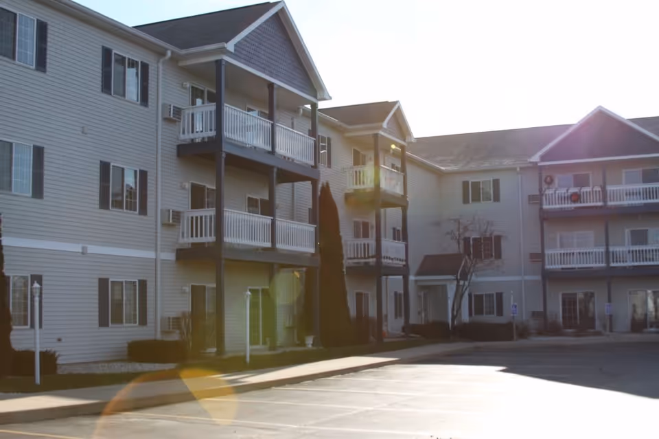 Exterior view of a three-story apartment building with balconies and multiple windows, under bright sunlight with lens flare effects. The building has beige siding and dark trim, with a parking lot in the foreground.