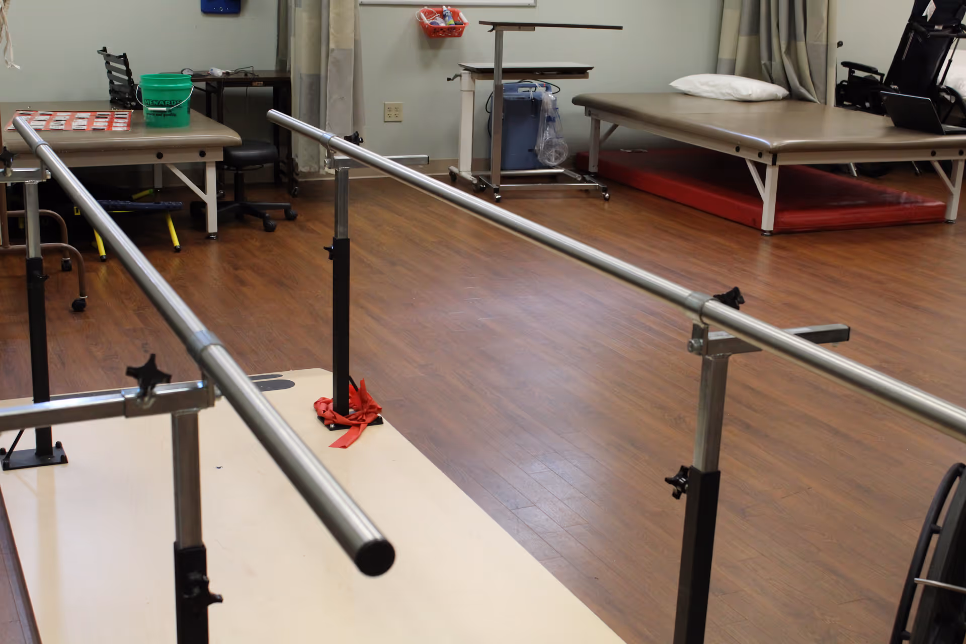 Physical therapy room with parallel bars for walking exercises, a treatment table with a pillow, a desk with a chair, and various therapy equipment on a wooden floor.