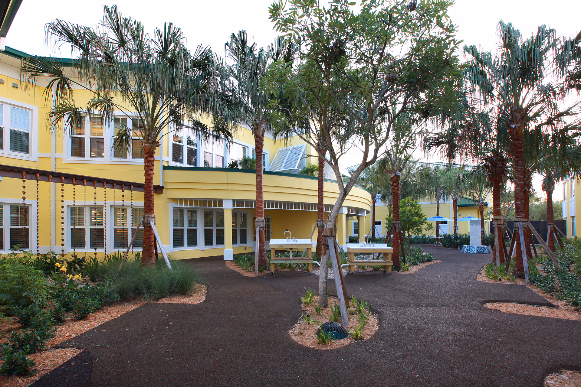 Outdoor courtyard area at The Cottage At Cypress Cove featuring a paved walking path, palm trees, benches, and a yellow two-story building with multiple windows in the background.