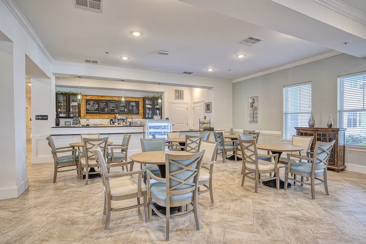 A bright and clean café area with several round tables and chairs arranged neatly. The café counter in the background has a menu board, display case, and pendant lights hanging from the ceiling. Large windows on the right side allow natural light to fill the room.