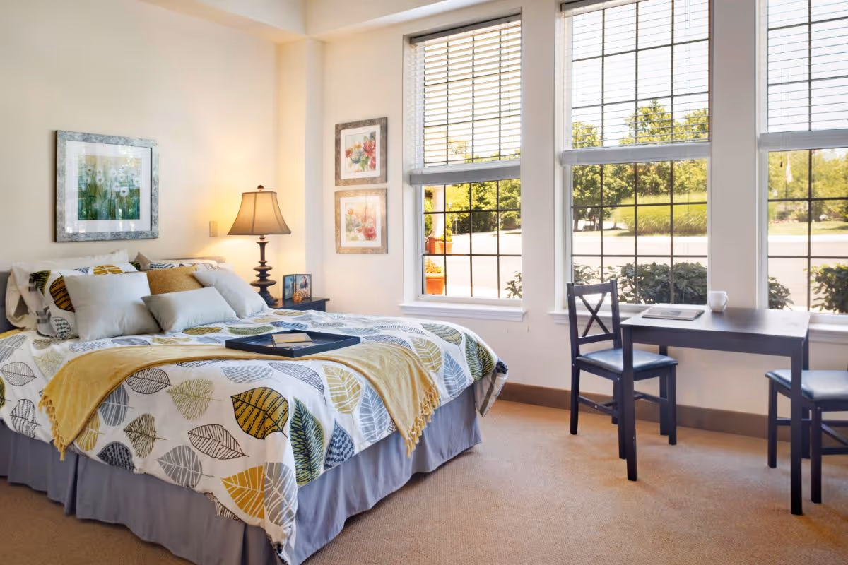 Sunlit bedroom with a made bed, bedside lamp, framed art, and a desk by large windows.