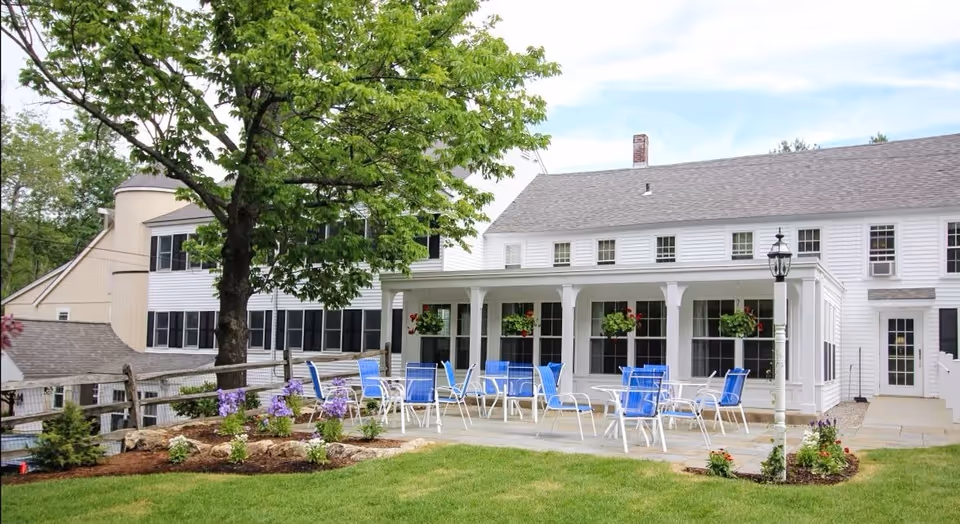 Outdoor patio area with multiple blue and white chairs and tables on a stone-paved surface, surrounded by green grass and flower beds. The patio is adjacent to a white building with large windows and hanging flower pots. A tree and a wooden fence are visible on the left side.