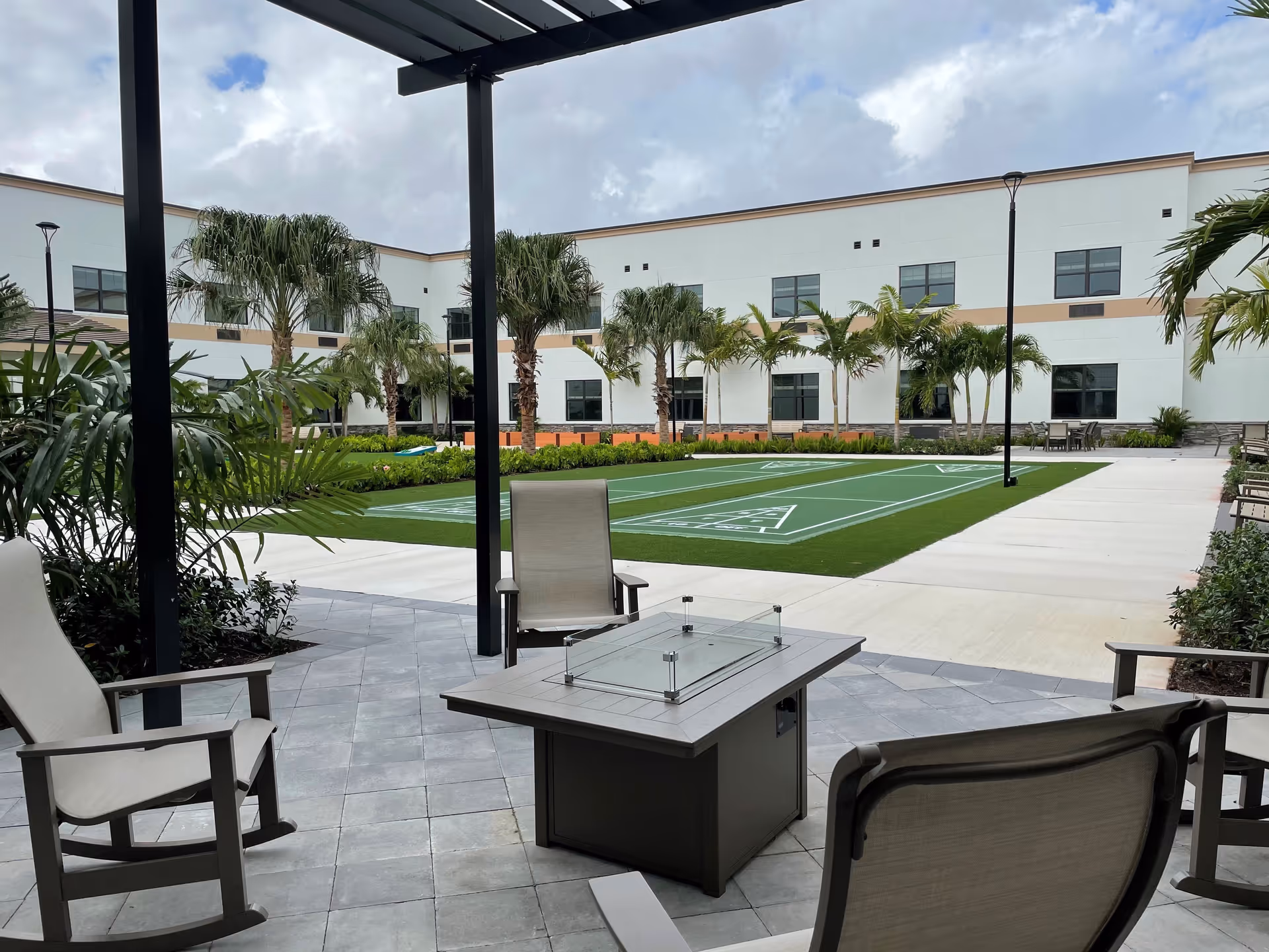 Outdoor patio area with several chairs surrounding a square fire pit table under a pergola. Beyond the patio is a green shuffleboard court with palm trees and a two-story white building in the background under a cloudy sky.