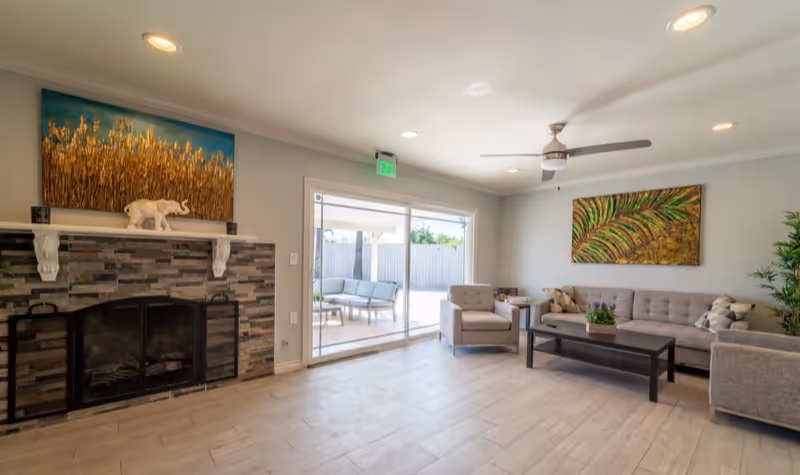 A bright living room with light-colored tiled flooring, a stone fireplace with a white mantle, and a decorative elephant figurine. The room has a gray sofa, two matching armchairs, a black coffee table with a small plant, and colorful artwork on the walls. A ceiling fan and recessed lighting are visible, and sliding glass doors lead to an outdoor patio area with additional seating.