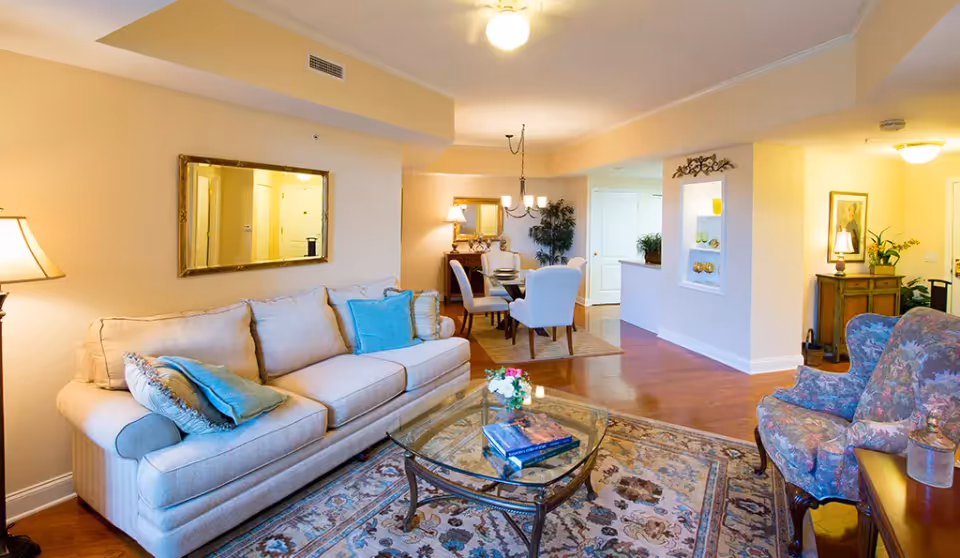 Bright furnished living room with a beige sofa, glass coffee table, patterned armchair, and an adjoining dining area.