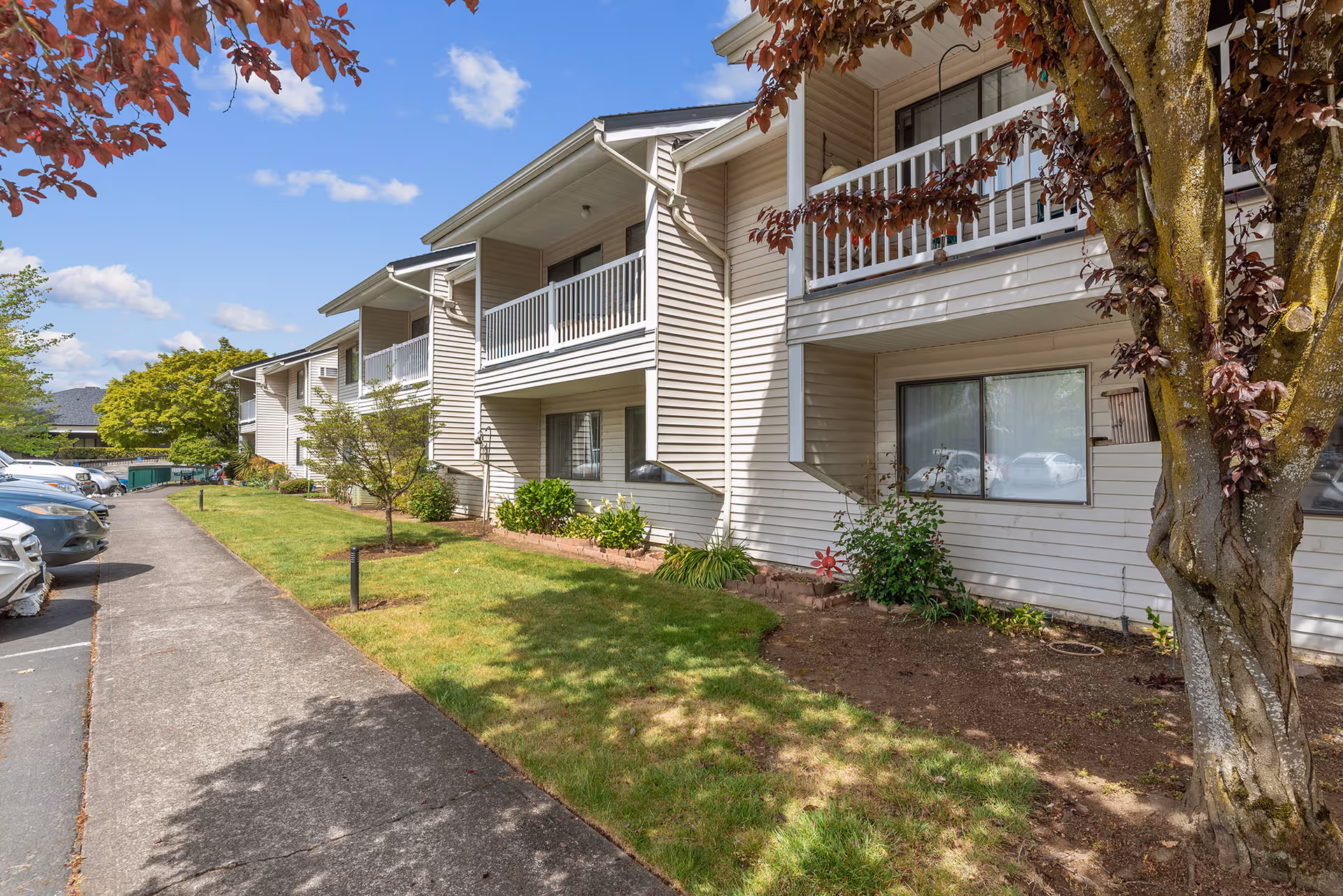 Exterior front of a two-story senior living apartment building with balconies, a lawn, walkway, and parked cars.