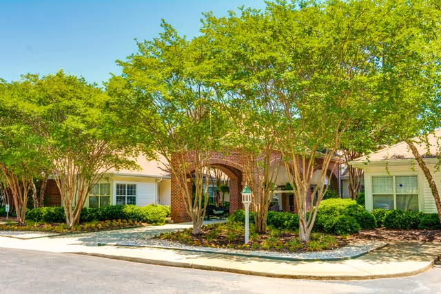 Exterior view of Harbison Shores Assisted Living and Memory Care facility showing a brick archway entrance surrounded by green trees and shrubs under a clear blue sky.