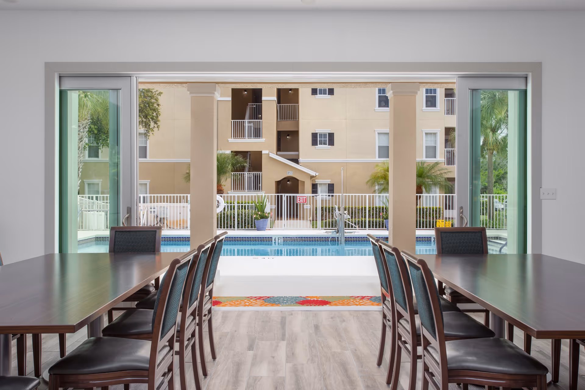 View from inside a dining room with two long wooden tables and chairs on either side, looking out through large sliding glass doors to an outdoor swimming pool and a multi-story beige building with balconies and palm trees.