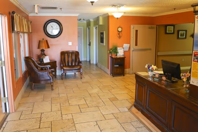 Reception area of a senior living facility with a wooden front desk on the right, a computer monitor, and small flower arrangements. On the left side, there are two brown leather chairs with a small table and lamp between them, a large wall clock above, and windows with curtains. The floor is tiled, and the walls are painted in warm tones of orange and green. A hallway extends in the background.