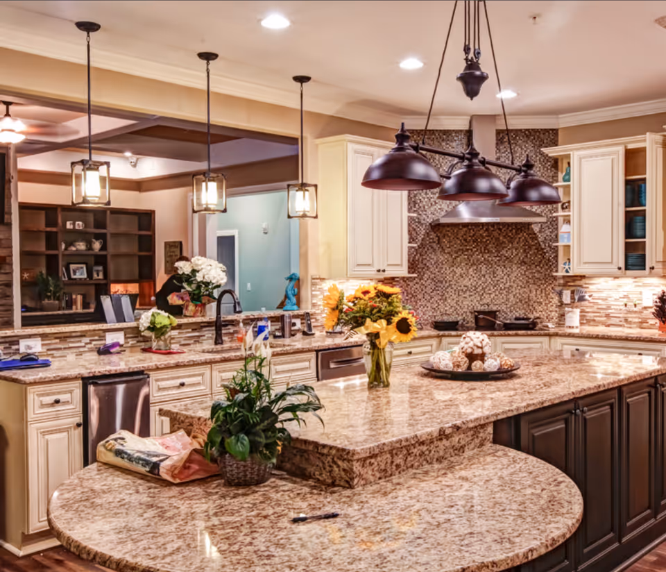 A spacious kitchen with granite countertops and a large island featuring a vase of sunflowers and decorative items. The kitchen has cream-colored cabinets, a mosaic tile backsplash, stainless steel appliances, and pendant lighting hanging from the ceiling. In the background, there is a view into a living area with shelves and a ceiling fan.