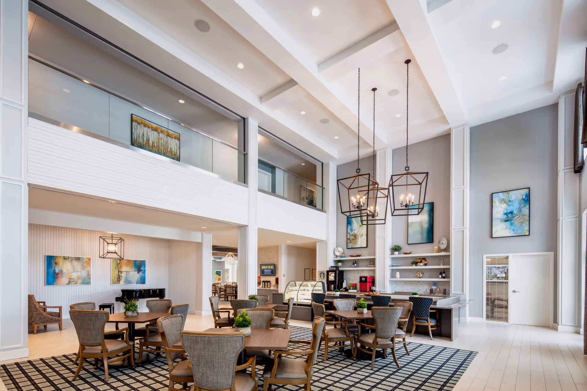 Bright two-story community dining and lounge area with round tables and chairs, high ceilings, pendant chandeliers, a café counter, and wall art.