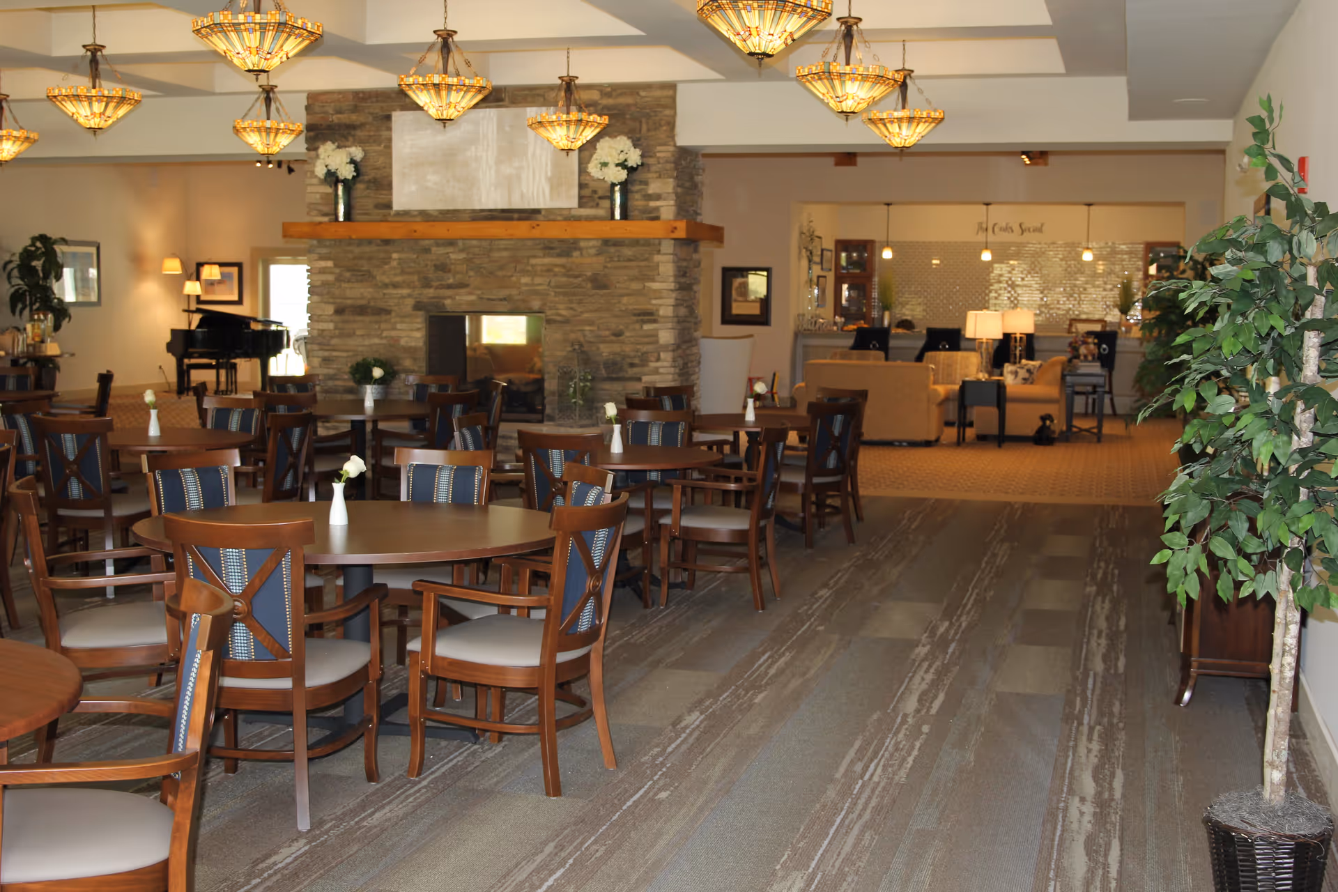 Spacious dining room with round wooden tables and chairs, a stone fireplace, chandeliers, and a lounge area in the background.