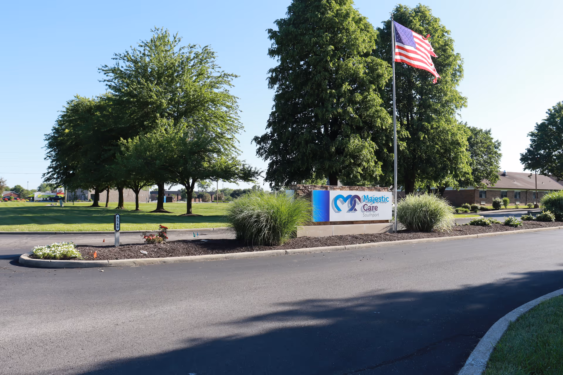 Entrance area of Majestic Care of Southport facility featuring a landscaped median with bushes and flowers, a sign displaying the facility's name and logo, an American flag on a tall flagpole, and several large trees in the background under a clear blue sky.