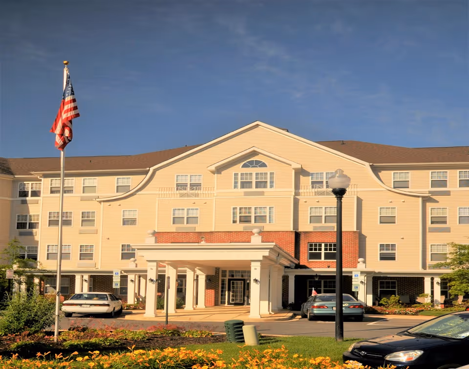Front exterior view of River Point Apartments, a multi-story building with beige siding and brick accents. The entrance features a covered portico with white columns. There are several cars parked in front, a flagpole with an American flag, a streetlamp, and landscaped flower beds in the foreground under a clear blue sky.