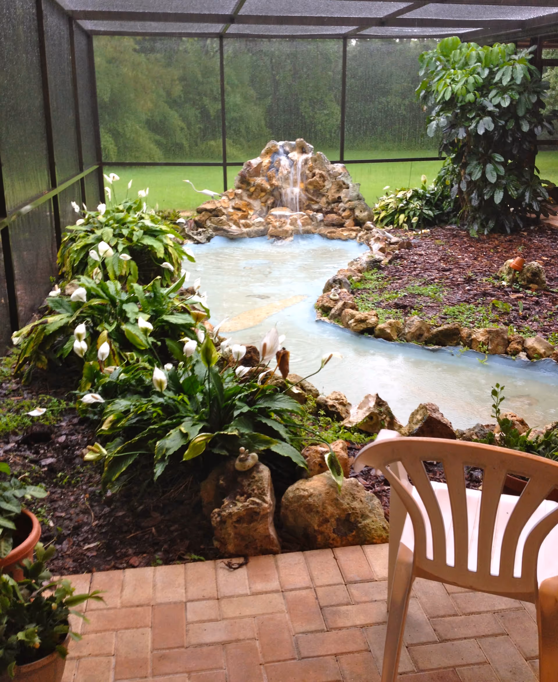 A screened-in outdoor garden area with a small artificial waterfall flowing into a shallow pond surrounded by rocks and green plants with white flowers. A plastic chair is partially visible in the foreground on a brick patio.