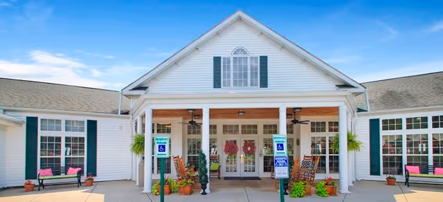 Front exterior view of Brookdale Reidsville facility featuring a white building with green shutters, a covered entrance supported by white columns, rocking chairs, potted plants, and reserved parking signs for handicapped access.