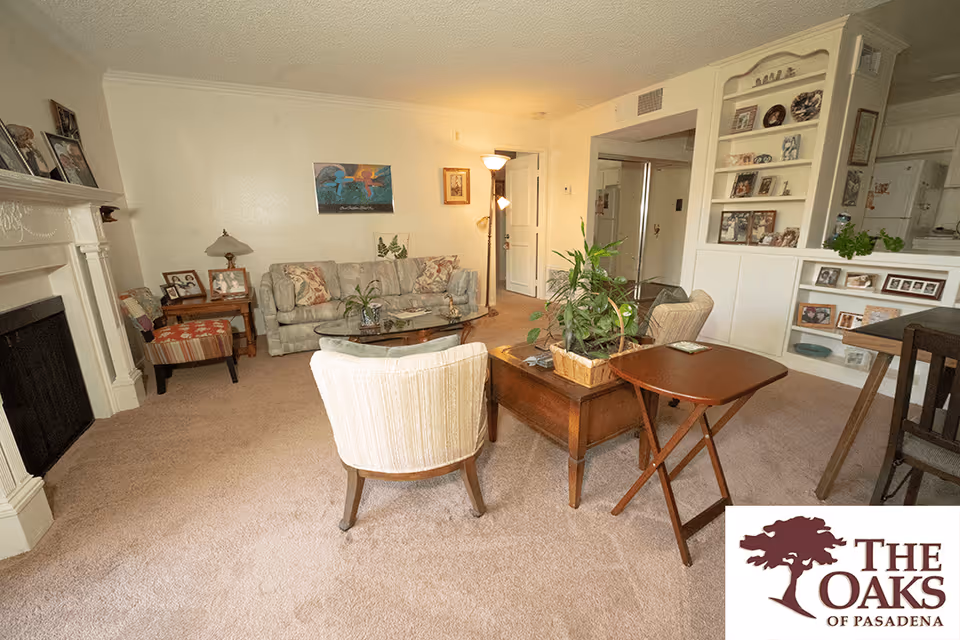 A cozy living room with beige carpet, a light-colored sofa with floral cushions, two armchairs, a glass coffee table, and a wooden side table with a plant. There is a fireplace on the left side with framed photos on the mantel. The room has built-in shelves with various decorative items and photos. A floor lamp stands near the door, and the kitchen is partially visible through an open wall space.