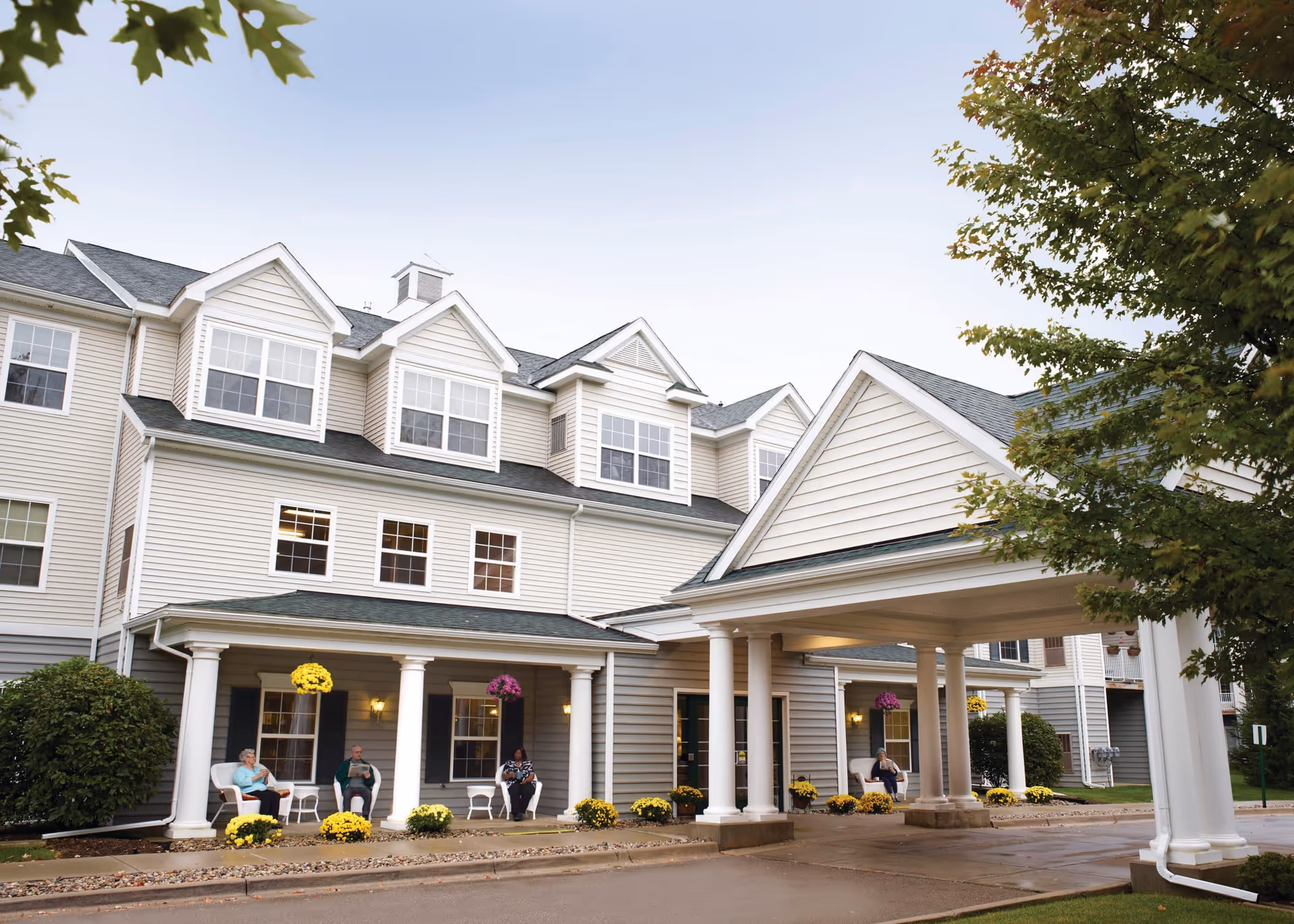 Exterior view of a senior living facility with a covered entrance supported by white columns. Three elderly people are sitting on chairs on the porch, surrounded by yellow and purple flowers. The building has multiple windows and a light-colored siding with a dark roof. Trees and bushes are visible around the building.