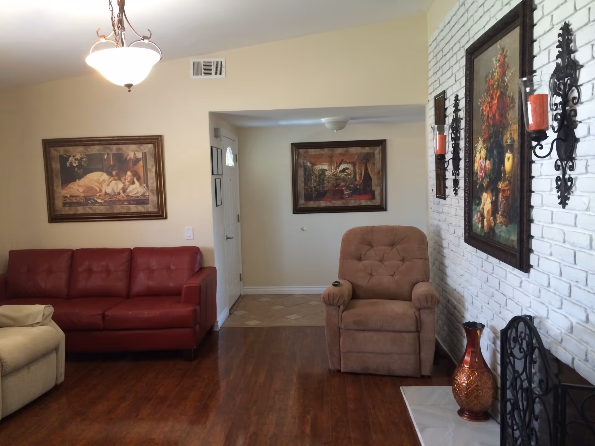 A cozy living room with a red leather sofa, a beige recliner, and a brown cushioned armchair. The room features wooden flooring, a white brick wall with floral paintings and decorative candle holders, and a hanging ceiling light. There is a doorway leading to another area with a framed picture on the wall.