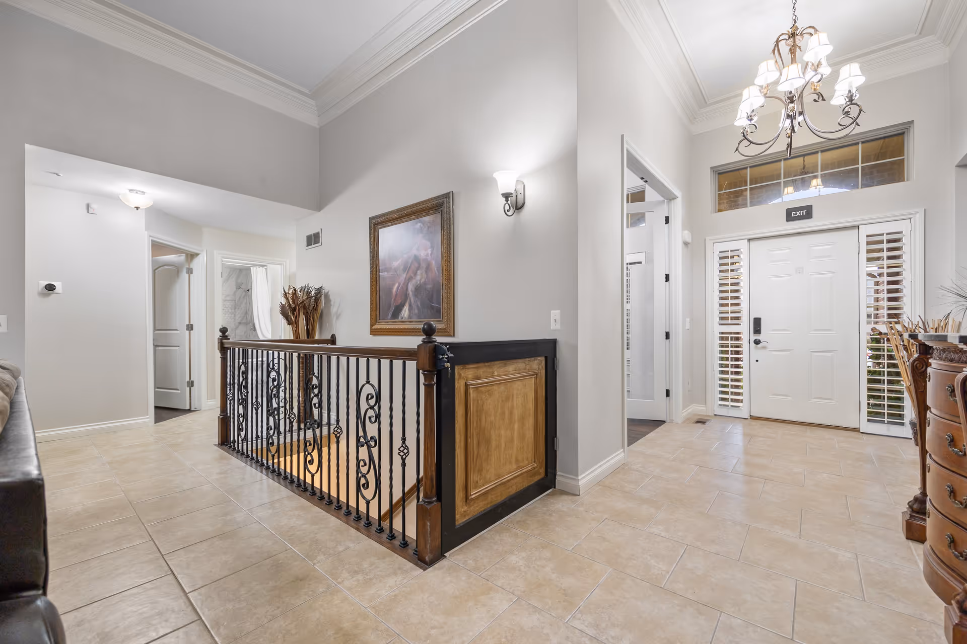 Interior view of a senior living facility hallway with beige tiled flooring, light gray walls, a decorative wrought iron railing around a stairwell, a framed painting on the wall, a chandelier hanging from the ceiling, and a white exit door with sidelights and an exit sign above it.