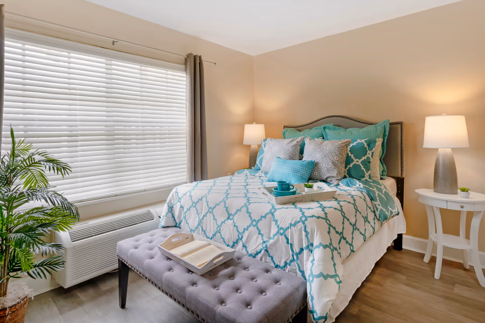 A cozy bedroom with a bed dressed in white and teal patterned bedding, multiple pillows, and a tray with a teal cup and small plant. There is a tufted gray bench at the foot of the bed with an open book on a tray. Two bedside tables with lamps flank the bed, and a large window with white blinds and gray curtains is on the left side. A potted plant is placed near the window.
