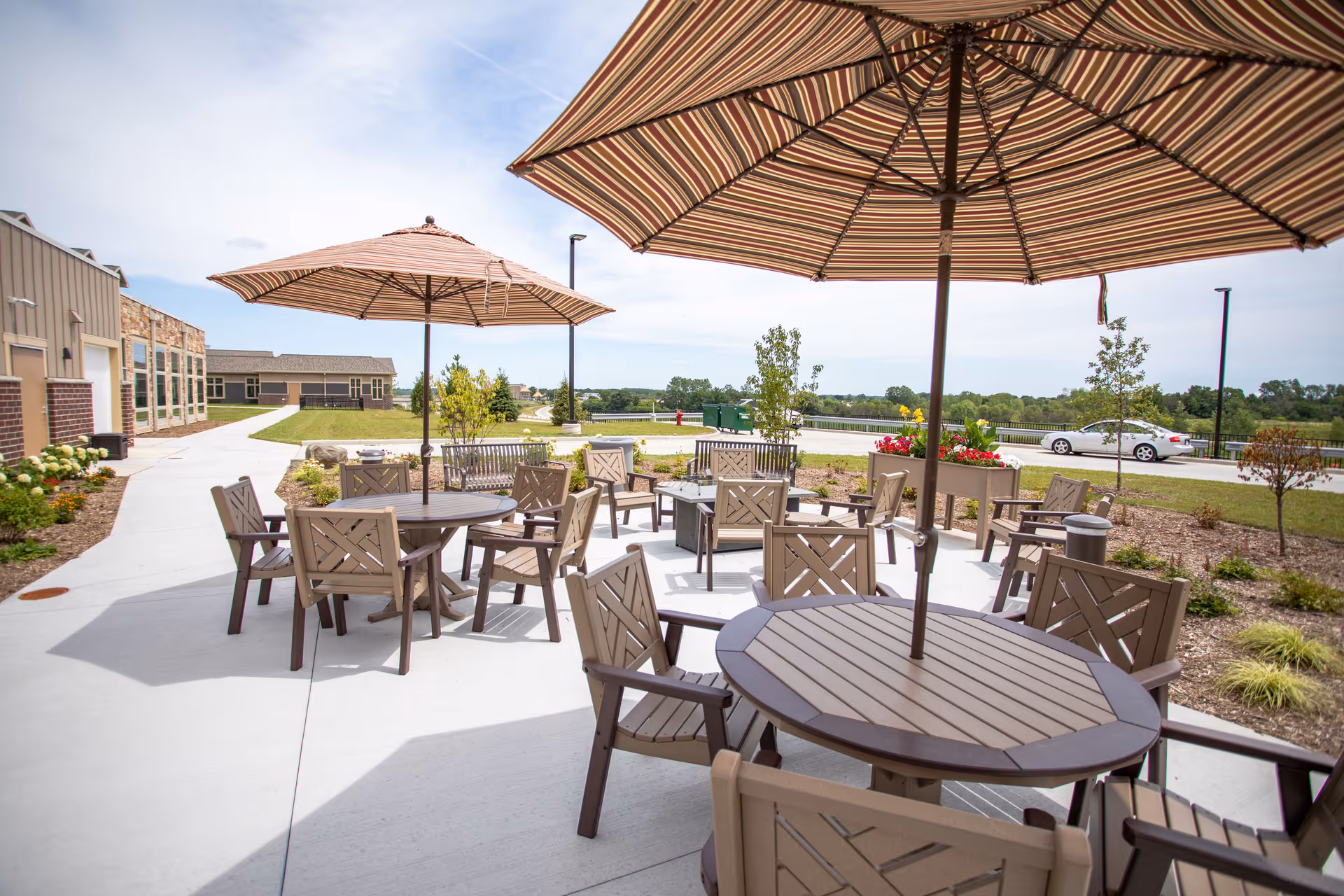 Outdoor patio area at New Perspective Senior Living | Franklin with multiple round tables and chairs under large striped umbrellas, surrounded by landscaped greenery and a clear sky.