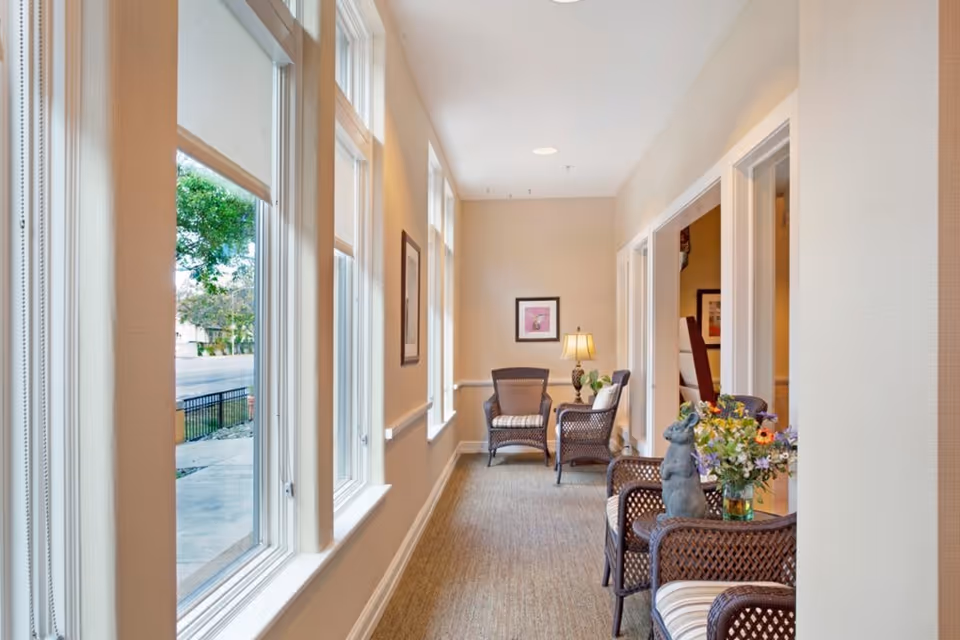 Sunlit interior hallway with wicker chairs, a side table with a lamp and flowers, and windows along one wall.