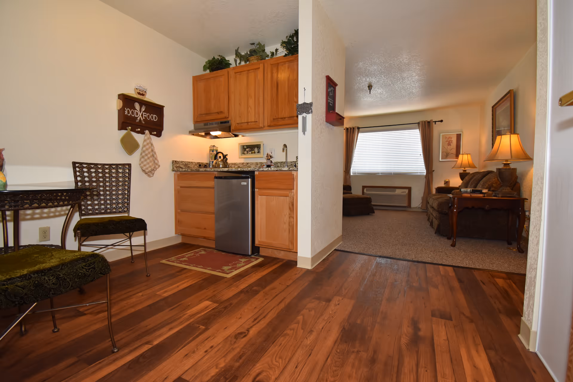 View of a small kitchen area with wooden cabinets, a mini refrigerator, and a granite countertop. To the left, there is a small dining table with two chairs. The kitchen opens into a living room with carpeted flooring, a sofa, side tables with lamps, and a window with curtains.