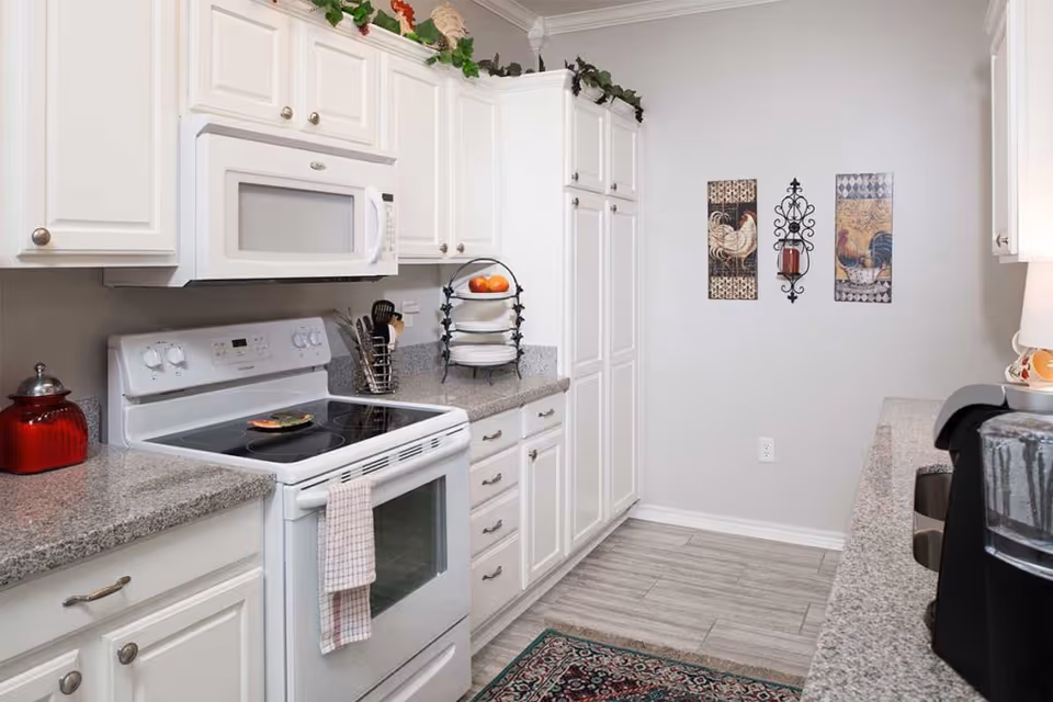 A clean and bright kitchen with white cabinets, a white electric stove with a microwave above it, granite countertops, and a decorative rug on the floor. The walls are light-colored with three decorative wall hangings featuring roosters and a candle holder. There are some kitchen utensils and a fruit stand on the counter.