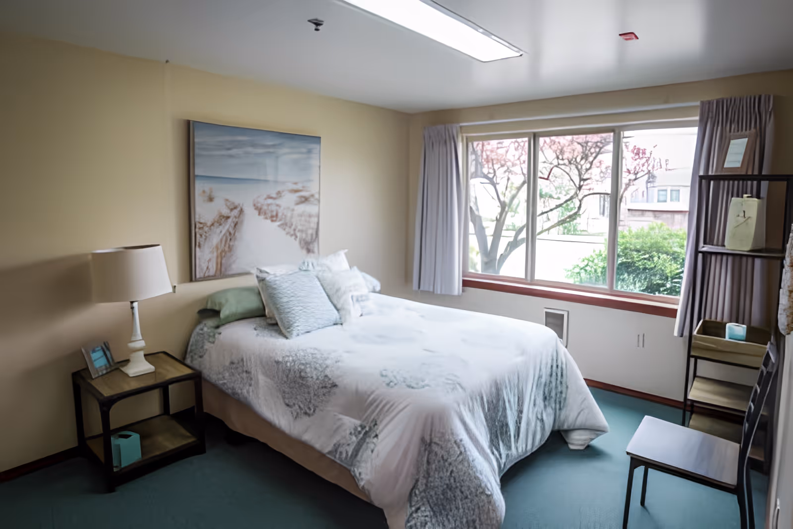A cozy bedroom with a bed covered in a light-colored comforter and multiple pillows. There is a nightstand with a lamp and a small picture frame on the left side of the bed. A large window with curtains lets in natural light, showing trees and greenery outside. On the right side, there is a shelving unit and a chair. The walls are painted beige and there is a framed picture hanging above the bed.