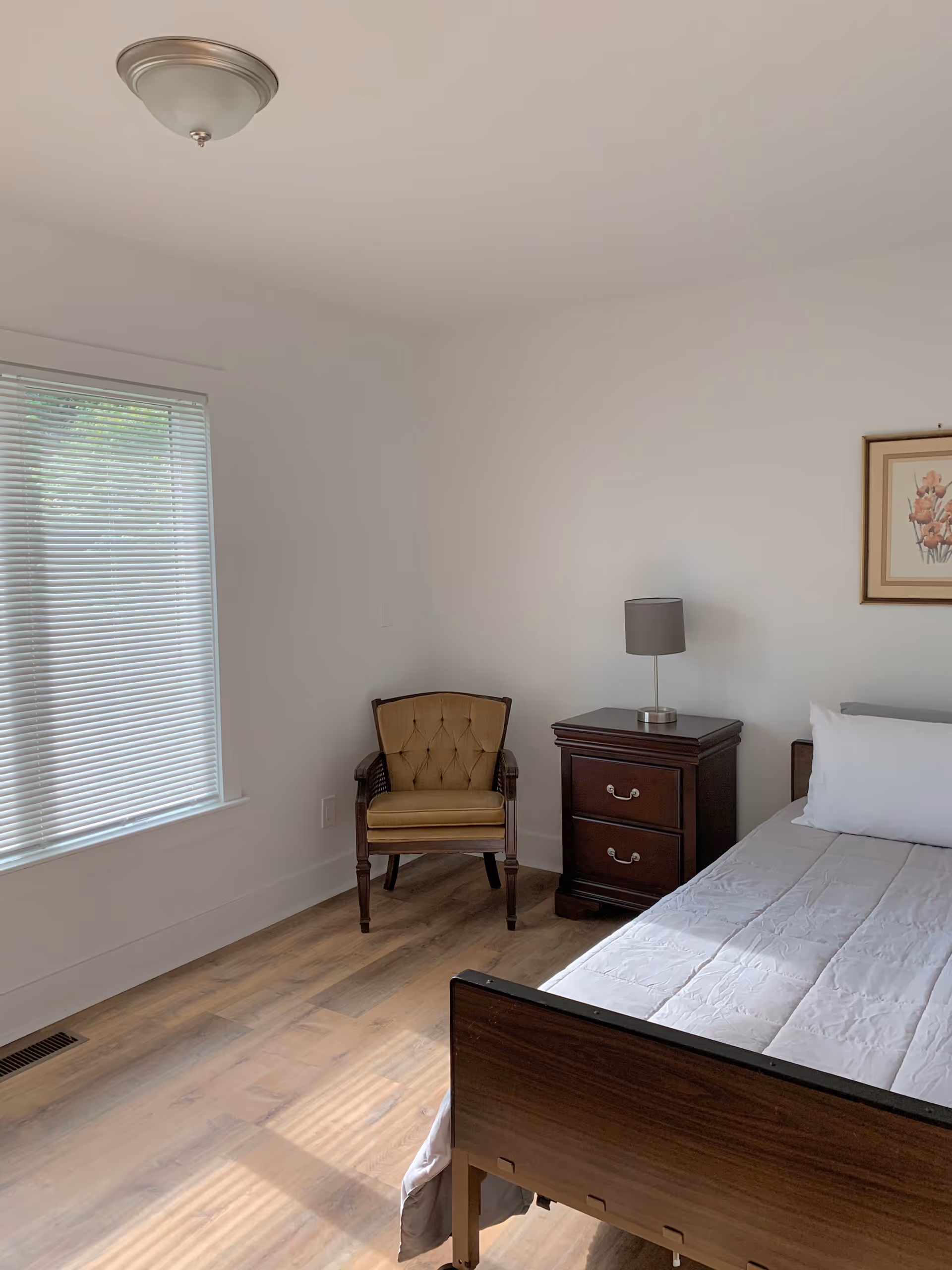 A simple bedroom with a single bed featuring a white pillow and light gray bedding. Next to the bed is a dark wooden nightstand with two drawers and a small gray lamp on top. A framed floral picture hangs on the wall above the nightstand. In the corner, there is a vintage-style upholstered chair with wooden arms and legs. The room has light-colored wooden flooring and a window with closed blinds allowing natural light to enter.