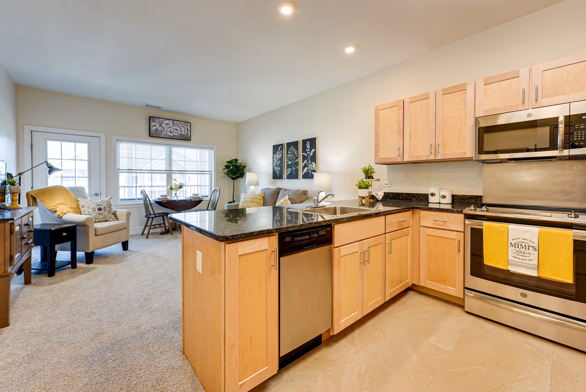 Bright and modern open-concept kitchen and living area in a senior living facility. The kitchen features light wood cabinets, a black granite countertop island with a built-in dishwasher, stainless steel microwave and oven, and a yellow towel hanging on the oven door. The living area has a beige armchair with a yellow throw, a small wooden dining table with chairs near a window, a couch with cushions, two table lamps, and wall art with botanical prints. A potted plant is placed near the window.