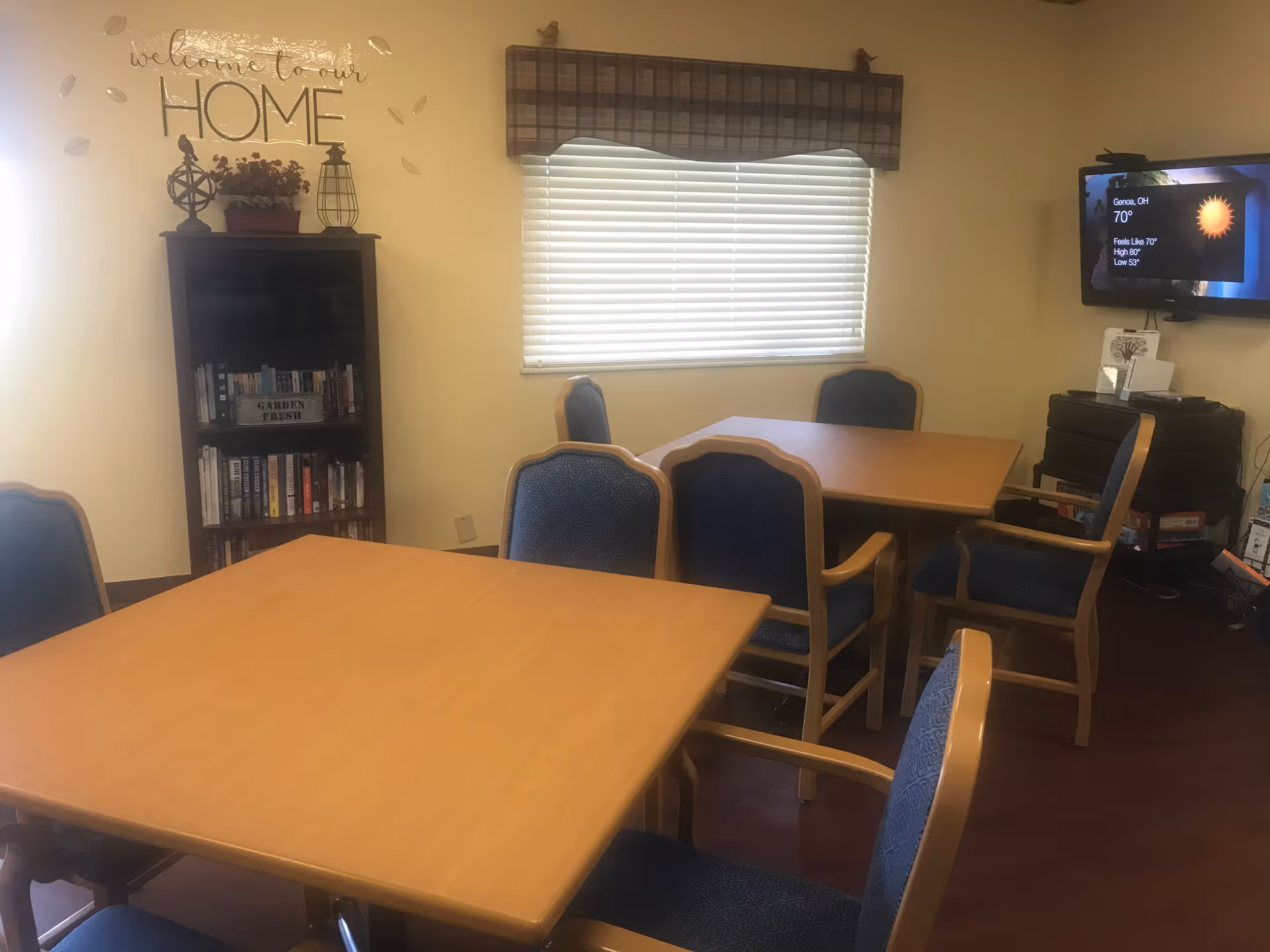 A cozy room with two wooden tables surrounded by blue cushioned chairs. A bookshelf filled with books is against the wall, above which is a decorative sign that reads 'welcome to our HOME'. A window with closed blinds and a plaid valance lets in natural light. A flat-screen TV mounted on the wall displays weather information for Genoa, OH.