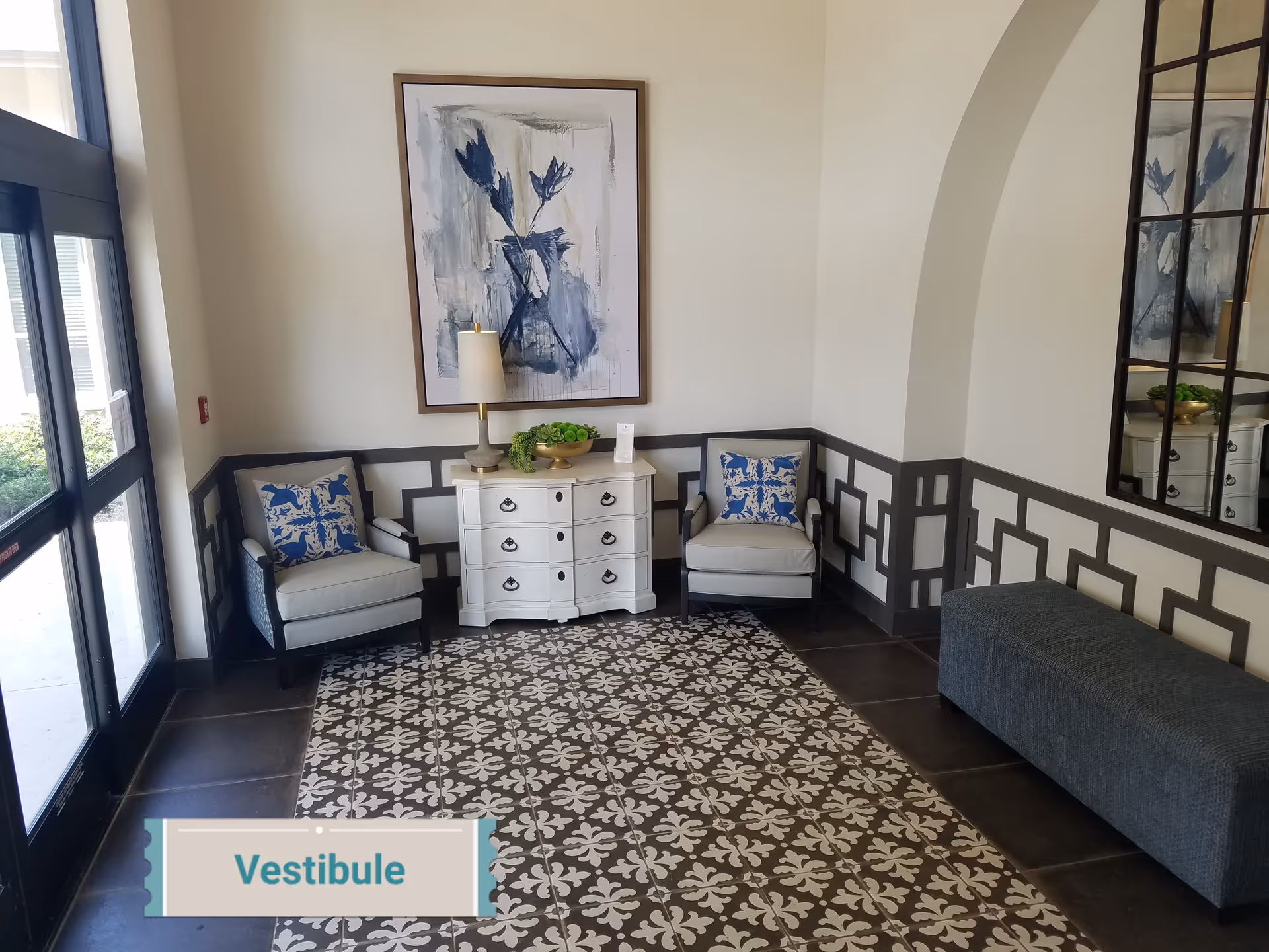 A small seating area in a vestibule with two cushioned armchairs featuring blue and white patterned pillows, a white chest of drawers with a lamp and decorative bowl on top, a large abstract painting with blue tones on the wall, a patterned tile floor, and a large windowed door letting in natural light.