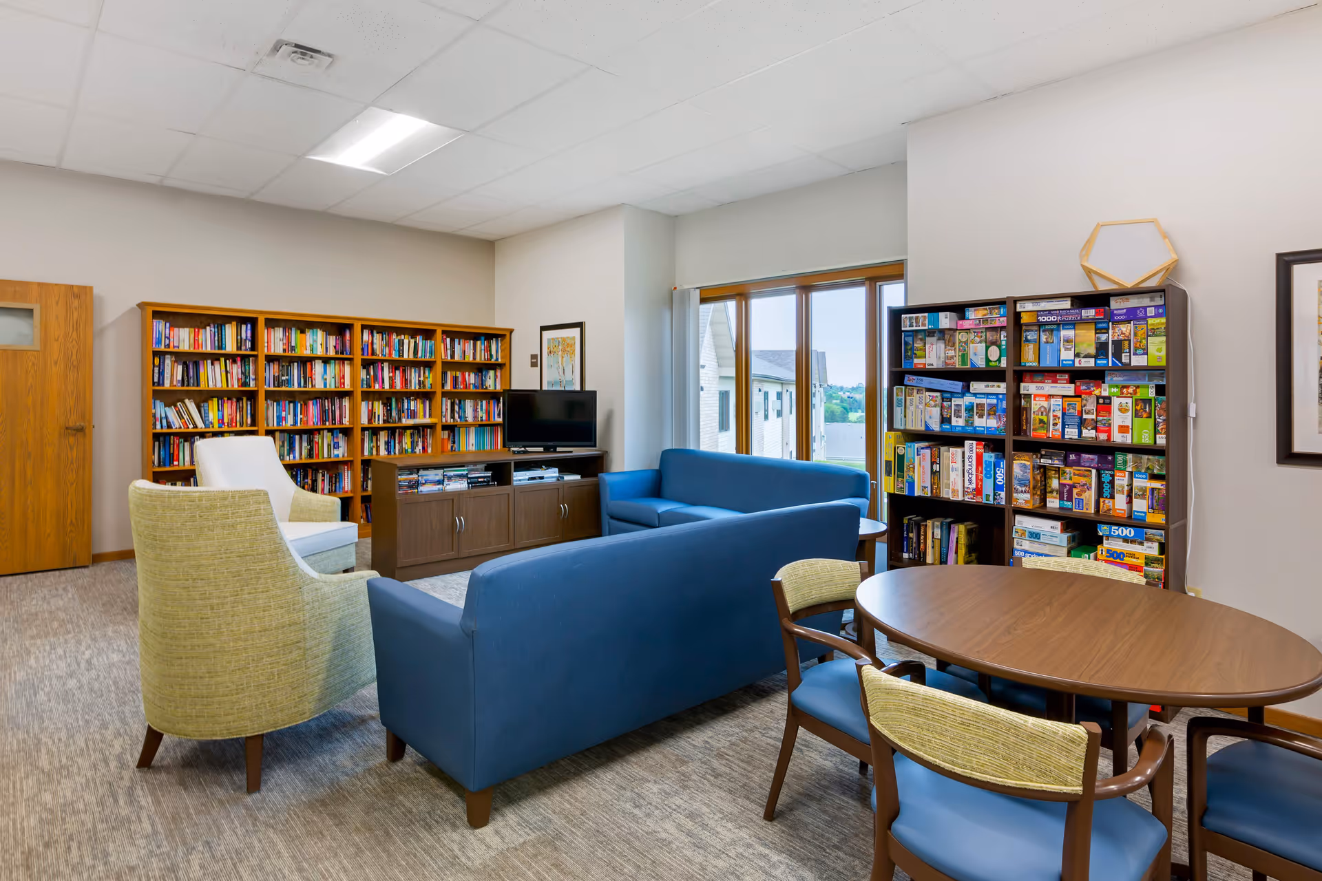 Community lounge with bookshelves, a game shelf, blue sofas and chairs, a TV, and a round table near a window.