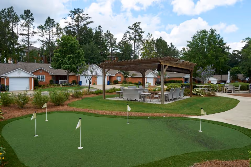 Outdoor area at Galleria Woods featuring a putting green with several small flags, a wooden pergola with seating underneath, surrounded by trees, shrubs, and residential buildings in the background under a partly cloudy sky.